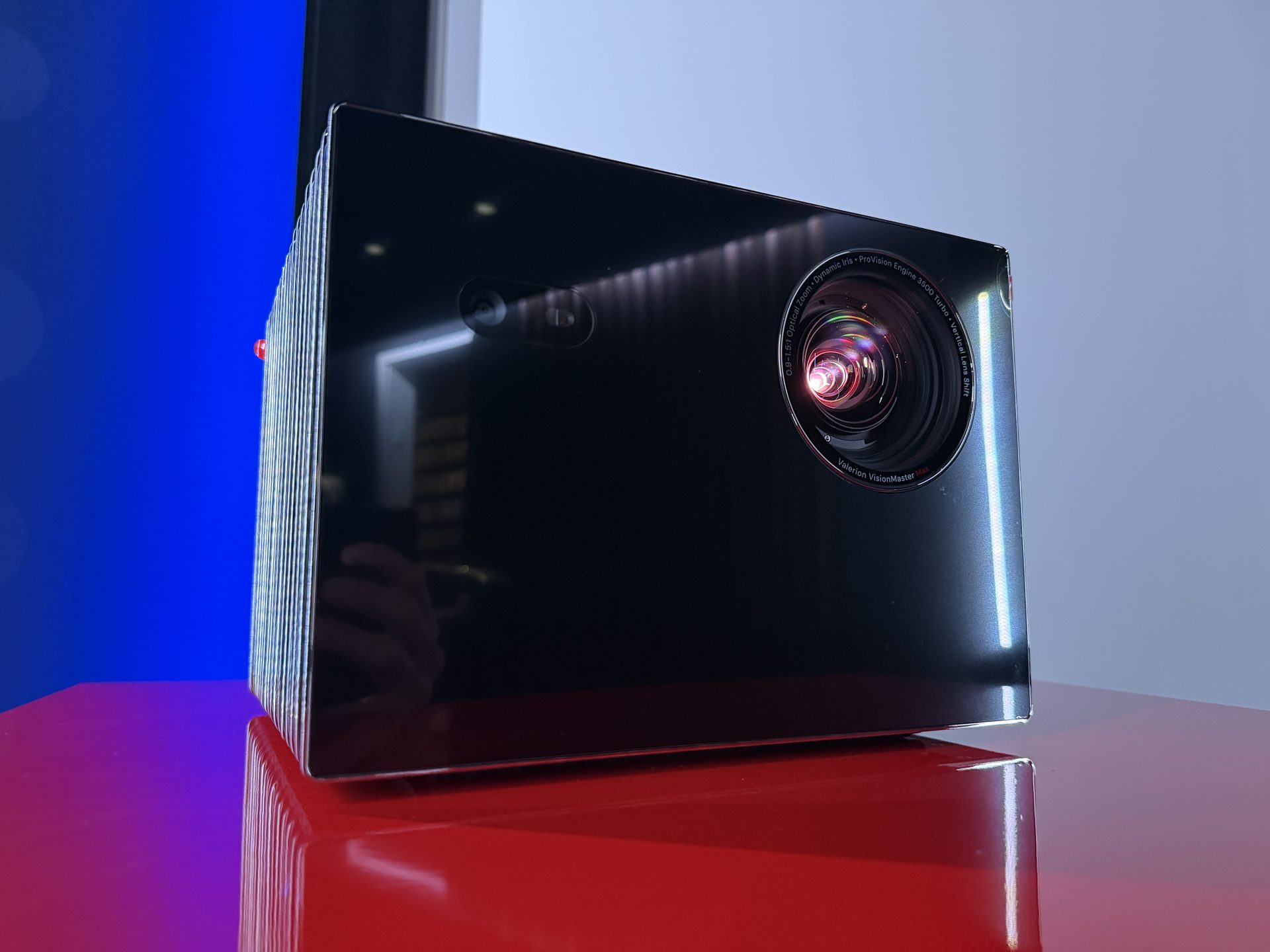 Der Projektor Valerion VisionMaster Max in Dreiviertelansicht auf einem rot lackierten Podest: schwarze Hochglanzfront, rechts das beleuchtete Objektiv, an der Seite feine Lamellen, im Hintergrund blaues und weißes Licht.