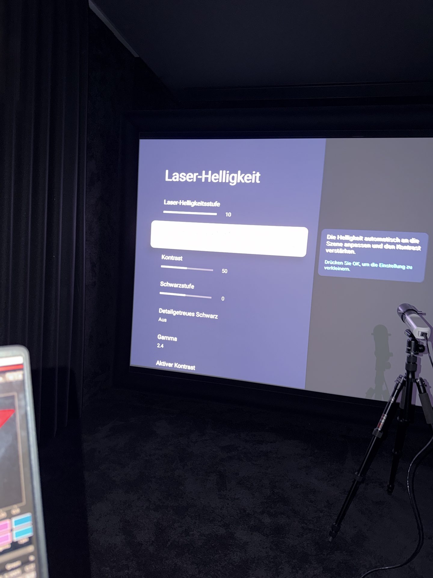 Projektormenü Laser-Helligkeit auf der Leinwand mit Laser-Helligkeitsstufe 10, Kontrast 50, Schwarzstufe 0, Detailgetreues Schwarz Aus und Gamma 2.4. Rechts steht der Messkopf auf einem Stativ, links im Anschnitt ein Laptop.