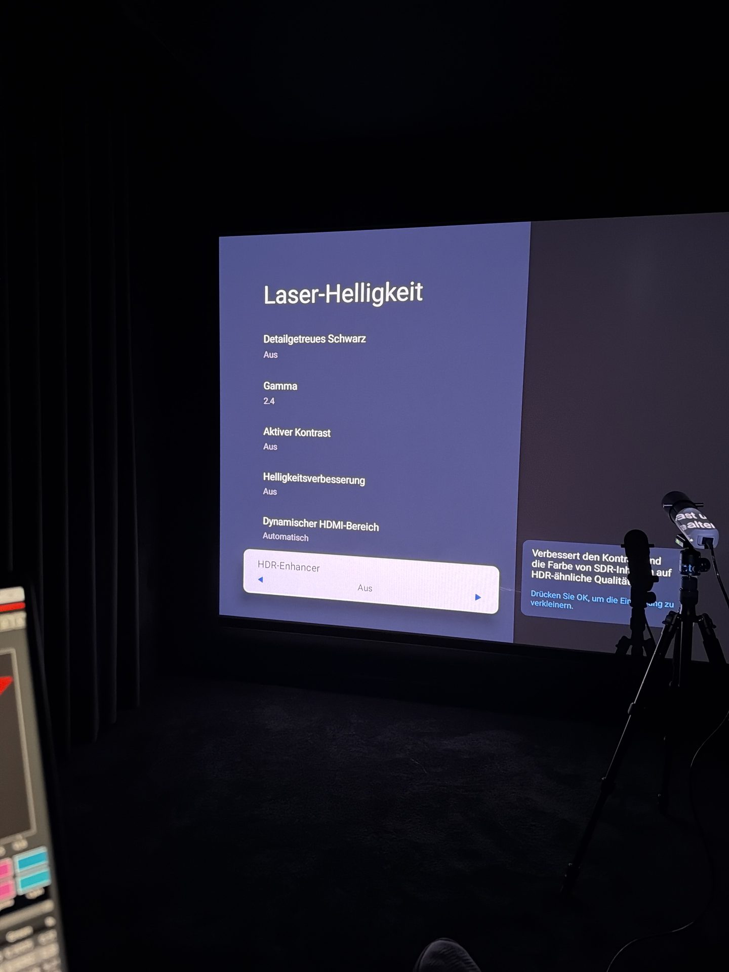 Projektormenü auf der Leinwand mit Gamma 2.4, Aktiver Kontrast Aus, Helligkeitsverbesserung Aus, Dynamischer HDMI-Bereich Automatisch und dem ausgewählten Punkt HDR-Enhancer Aus. Davor die Silhouette des Messkopfs auf dem Stativ.